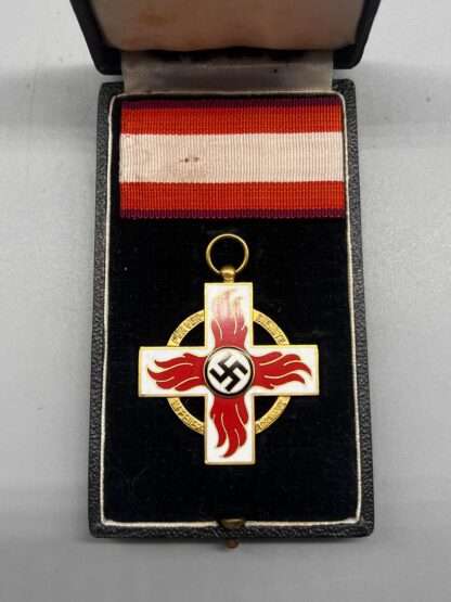 A Fire Brigade Honour Cross 1st Class Medal with presentation case. The medal depicts a Geneva-style cross in white enamel with sylised red enamel flames with a mobile swastika in the centre in black enamel, which sits in the presentation box with period ribbon.