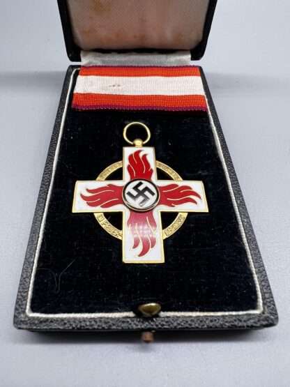 A Fire Brigade Honour Cross 1st Class Medal with presentation case. The medal depicts a Geneva-style cross in white enamel with sylised red enamel flames with a mobile swastika in the centre in black enamel, which sits in the presentation box with period ribbon.
