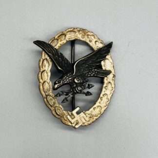 A photo of Luftwaffe Air Gunner & Flight Engineers Badge, which includes an oval shaped wreath of oak and laurel leafs with a swastika at the base. In the centre includes a national eagle "flying from left to right in a downwards direction"; clutched in its claws were two crossed arrow-head lightning bolts.
