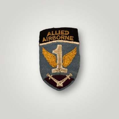 A first allied airborne formation patch, the cloth badge depicts a light blue shield at the top of which is a black band with the words 'Allied Airborne' in yellow; below, a white figure '1' with yellow wings above a pair of crossed gladiator swords, point down, on a purple-red background. The badge has no damage or repairs in good condition.