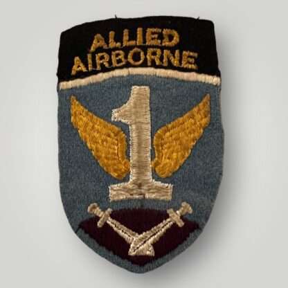 A first allied airborne formation patch, the cloth badge depicts a light blue shield at the top of which is a black band with the words 'Allied Airborne' in yellow; below, a white figure '1' with yellow wings above a pair of crossed gladiator swords, point down, on a purple-red background. The badge has no damage or repairs in good condition.