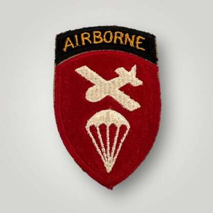 A US WW2 Airborne Command Patch, machine embroidered. The insignia depicts a red shield with a glider and parachute in white thread, with a scroll above with the inscription "Airborne" embroidered in orange thread on black backing.