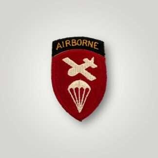 A US WW2 Airborne Command Patch, machine embroidered. The insignia depicts a red shield with a glider and parachute in white thread, with a scroll above with the inscription "Airborne" embroidered in orange thread on black backing.