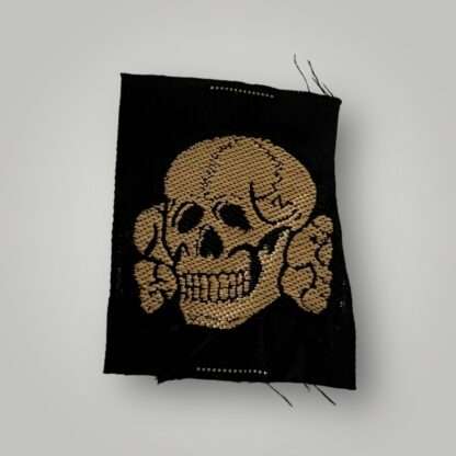 An original unissued Waffen-SS tropical cap skull insignia, flat wire machine embroidered BeVo construction in tan coloured thread on black coloured rayon.