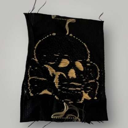 Reverse image of an Waffen-SS tropical cap skull insignia, flat wire machine embroidered BeVo construction in tan coloured thread on black coloured rayon.
