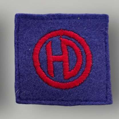 An original British WW2 51st Highland Division formation badge, machine embroiderd depicting the letters HD conjoined in red within a red circle on a blue woollen backing.