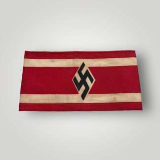 A Hitler Youth Student Bund Armband. The armband is constructed in red cotton with two white stripes along the edges. In the middle, there is a black swastika inside a white diamond, which is a typical symbol of the Hitler Youth.