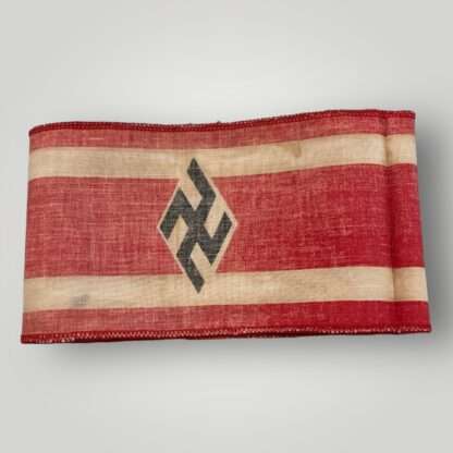 Reverse side of a Hitler Youth Student Bund Armband. The armband is constructed in red cotton with two white stripes along the edges. In the middle, there is a black swastika inside a white diamond, which is a typical symbol of the Hitler Youth