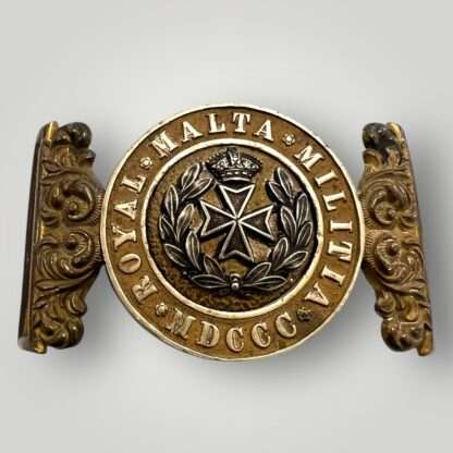A Royal Malta Militia belt buckle 1889 – 1903, constructed in brass and white metal. The buckle is a two-part design with ornate, floral filigree borders on the outside. A round, recessed center embossed with "Royal Malta Militia, MDCCC".