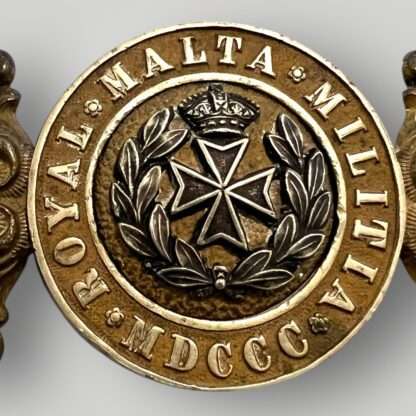 A Royal Malta Militia belt buckle 1889 – 1903, constructed in brass and white metal. The buckle is a two-part design with ornate, floral filigree borders on the outside. A round, recessed center embossed with "Royal Malta Militia, MDCCC".