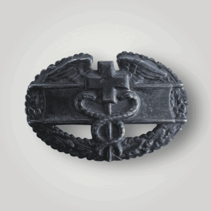 A rare United States WW2 Combat Medic Badge. The oval-shaped badge features a cross flanked by wings on either side, with a rod below entwined by two serpents, superimposed over a stretcher and encircled by a wreath of oak leaves.