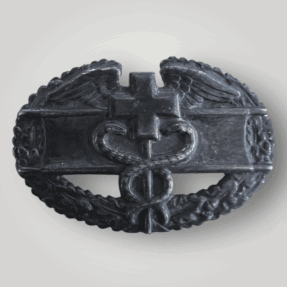 A rare United States WW2 Combat Medic Badge. The oval-shaped badge features a cross flanked by wings on either side, with a rod below entwined by two serpents, superimposed over a stretcher and encircled by a wreath of oak leaves.