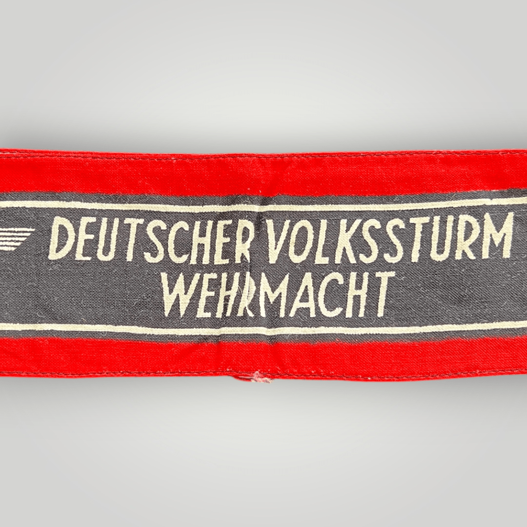 This is an original Deutscher Volkssturm Wehrmacht armband made of screen-printed cotton. It features a central black band with white lettering and Wehrmacht Eagles, bordered by two white lines and red edges.