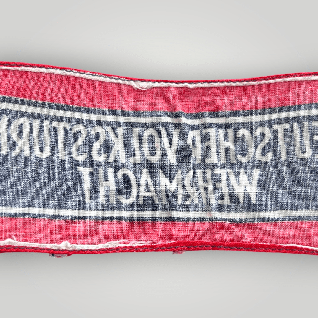 This is an original Deutscher Volkssturm Wehrmacht armband made of screen-printed cotton. It features a central black band with white lettering and Wehrmacht Eagles, bordered by two white lines and red edges.
