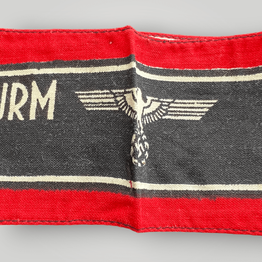 This is an original Deutscher Volkssturm Wehrmacht armband made of screen-printed cotton. It features a central black band with white lettering and Wehrmacht Eagles, bordered by two white lines and red edges.