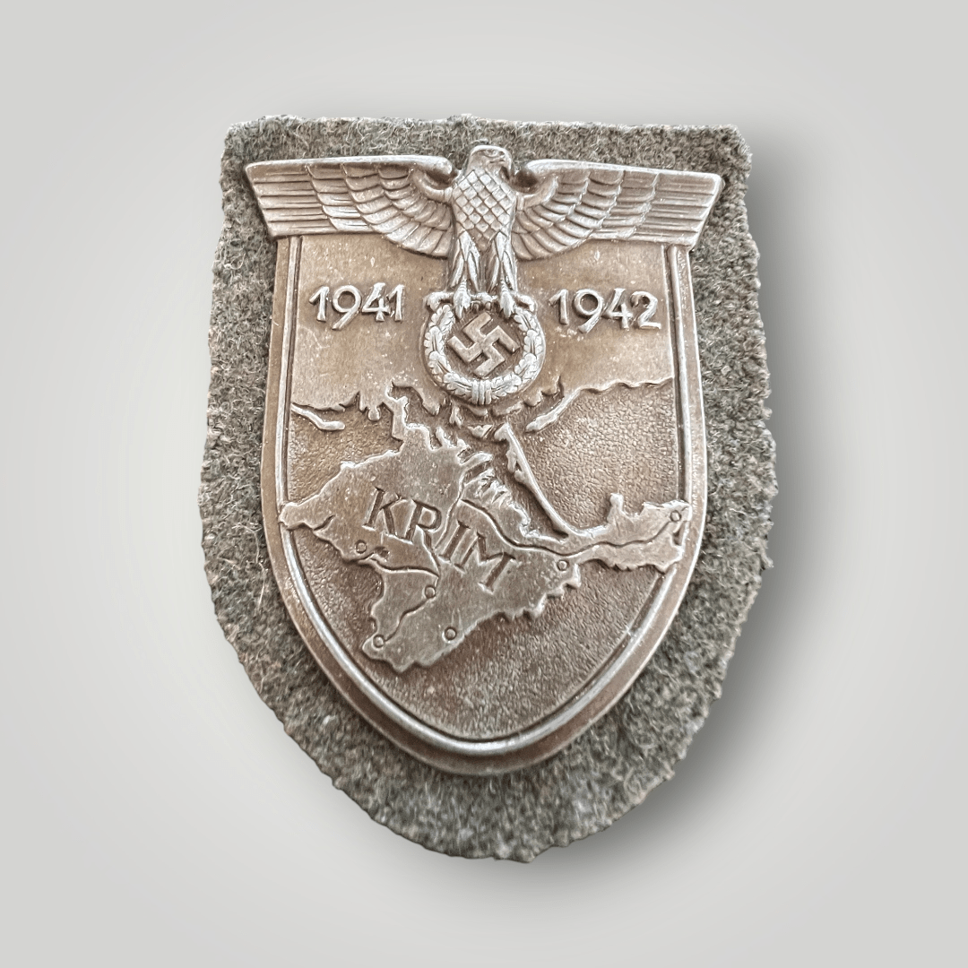 An original Heer Krim Campaign Shield, constructed in pressed steel, magnetic, with a bronze finish. The obverse of the decortaion depicts a raised map of Crimea and southern Ukraine, topped by a Wehrmacht-style German national eagle clutching a wreathed mobile swastika, flanked by dates “1941-1942”, affixed to a field-grey wool patch.