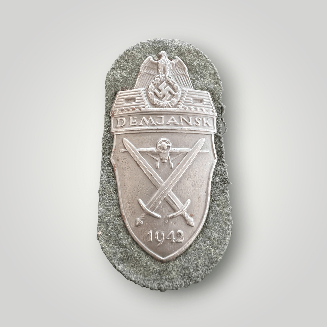 An original Heer Demjansk shield, die-struck with silver wash. The obverse depicts the national eagle clutching a wreathed mobile swastika, with a log pillbox on either side, with the inscription above “DEMJANSK”, with a frontal image of a prop plane below, with crossed swords with the date below “1942”, mounted on field-grey woolen background.