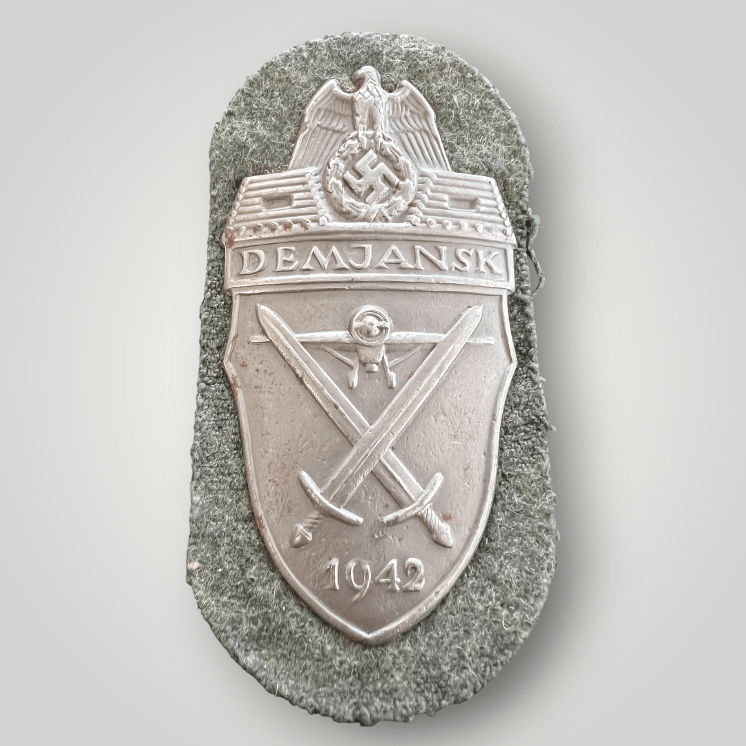 An original Heer Demjansk shield, die-struck with silver wash. The obverse depicts the national eagle clutching a wreathed mobile swastika, with a log pillbox on either side, with the inscription above “DEMJANSK”, with a frontal image of a prop plane below, with crossed swords with the date below “1942”, mounted on field-grey woolen background.