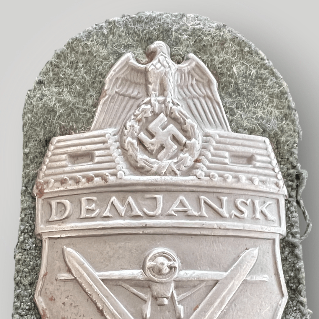 An original Heer Demjansk shield, die-struck with silver wash. The obverse depicts the national eagle clutching a wreathed mobile swastika, with a log pillbox on either side, with the inscription above “DEMJANSK”, with a frontal image of a prop plane below, with crossed swords with the date below “1942”, mounted on field-grey woolen background.