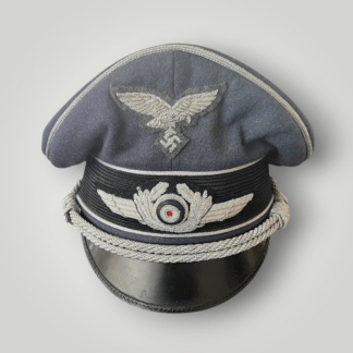 Luftwaffe Officer's Visor Cap By Erel, constructed in field grey blue wool pipped with silver aluminium pipping around the ridge of the cap and above and below the central band. The visor cap insignia includes hand embroidered silver aluminium cap eagle, and winged cockade.