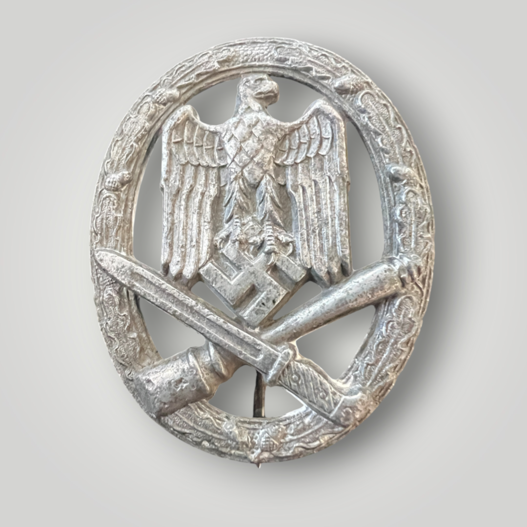 An original General Assault Badge by Assmann, constructed in zinc using a process called liquid-mold casting. The badge features an oval oak leaf wreath with acorns, with a German national eagle facing right, clutching a swastika, positioned above a crossed bayonet and a stick hand grenade.