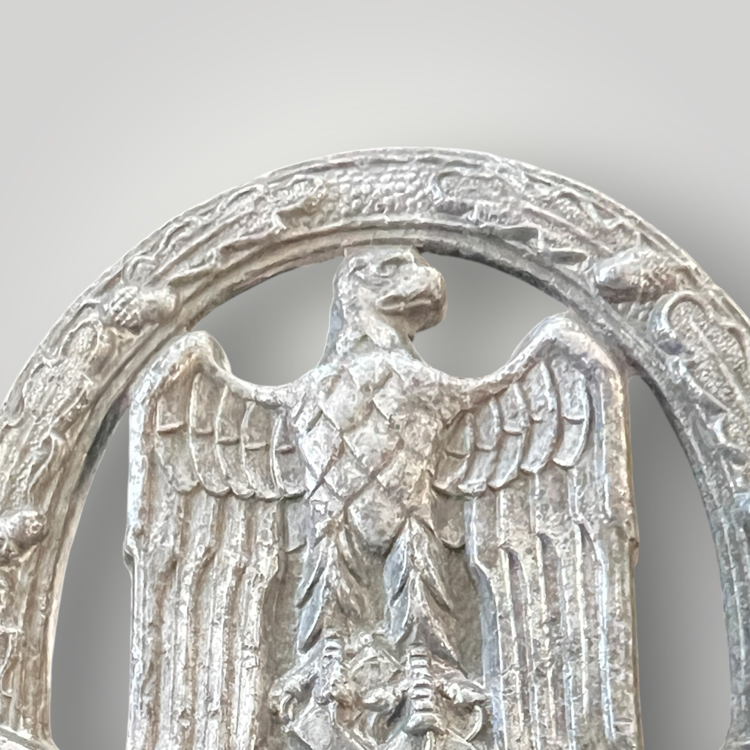 An original General Assault Badge by Assmann, constructed in zinc using a process called liquid-mold casting. The badge features an oval oak leaf wreath with acorns, with a German national eagle facing right, clutching a swastika, positioned above a crossed bayonet and a stick hand grenade.
